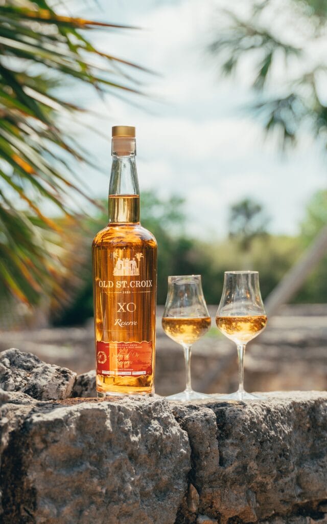 bottle of XO Amdre d'Or and two nosing lasses standing on rough stone wall against tropical background