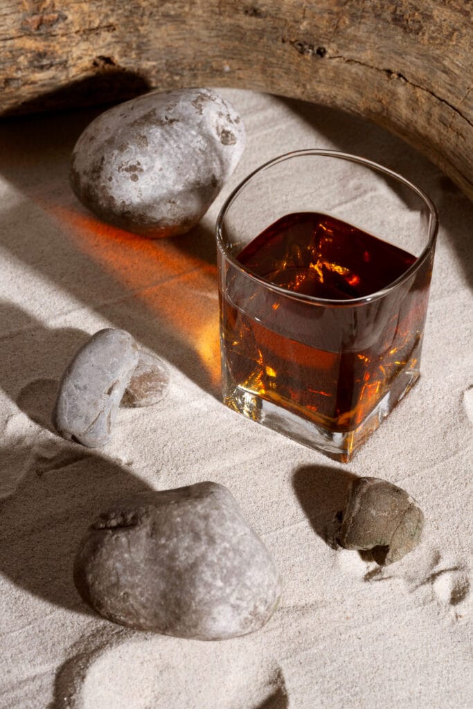 a whisky glass with a brown fluid sitting on a sandy floor